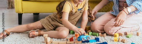 cropped view of babysitter near child playing with toy car near multicolored blocks on floor, horizontal image