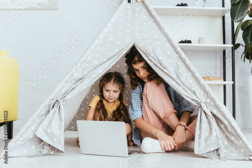 Fotografie young nanny sitting with crossed legs near cute kid using laptop in toy wigwam