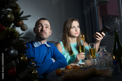 Bored or quarreled young man and woman sitting at holiday table and watching tv at Christmas night