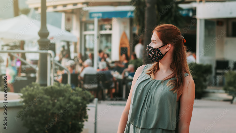 Fototapeta premium Woman in protective fashionable face mask in a touristic place during covid new normal with crowded street cafe in background, sunset time