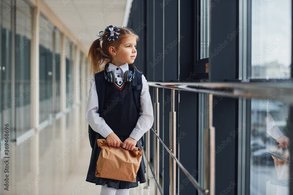 Fototapeta premium Young little school girl in uniform standing in hallway with package of dinner in hands