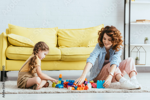 smiling babysitter and cute kid playing with building blocks while sitting on floor near yellow sofa