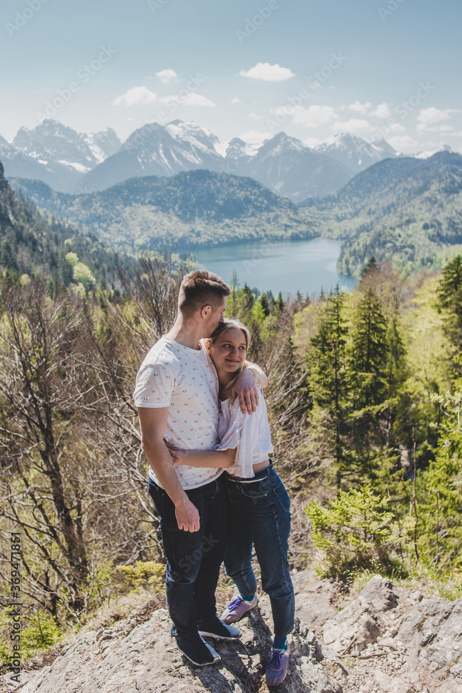 Lovely caucasian couple in white T-shirts stand on the top & embrace on background of Alpsee lake, green forest, rock mountains with snow peaks & blue sky. Bavaria. Germany