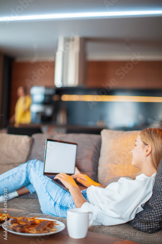 Pretty teen girl lying on sofa with blank screen laptop watching TV series with sweets and tea. Cozy home environment