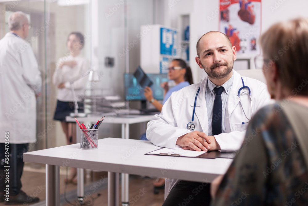 Fototapeta premium Young doctor looking worried at elderly aged patient during medical examination in hospital office. Young sick woman talking with mature medic on clinic corridor about treatment for her illness and