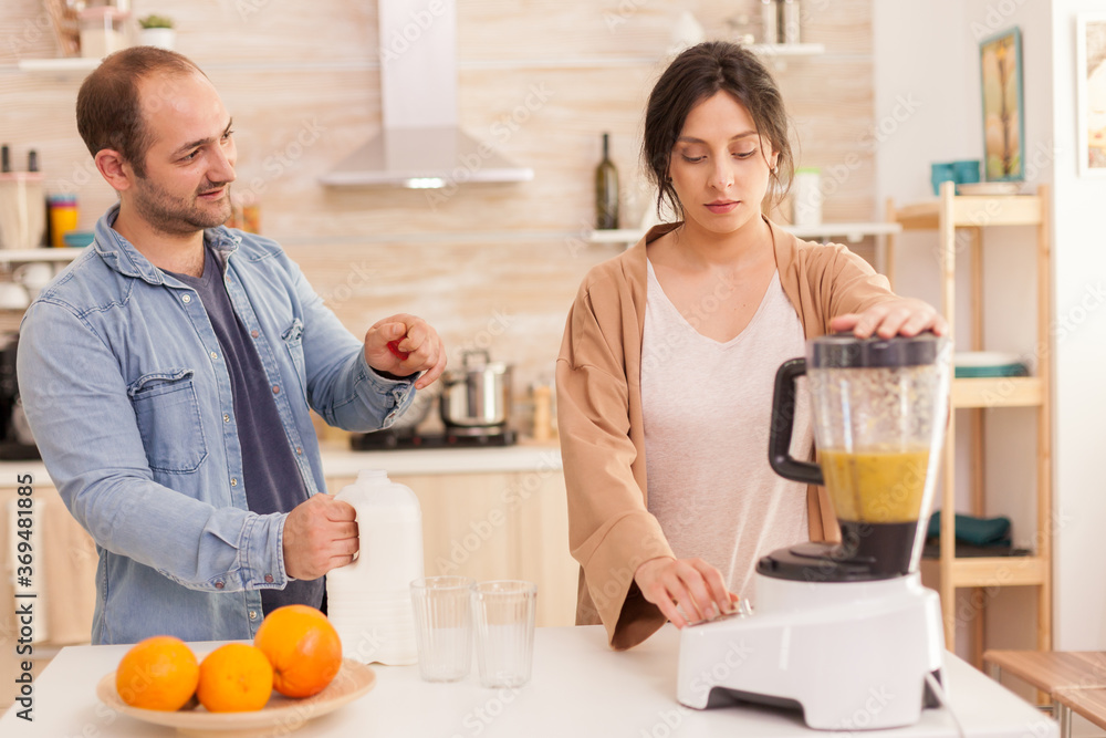 Woman mixing fruits in smoothie maker while husband opens milk bottle. Healthy carefree and cheerful lifestyle, eating diet and preparing breakfast in cozy sunny morning
