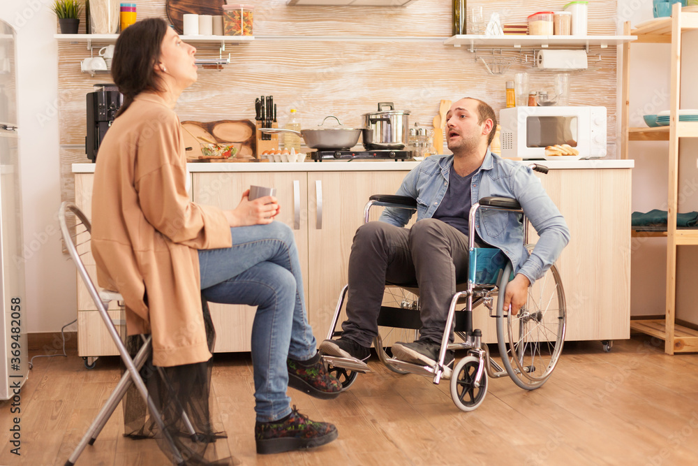 Angry man in wheelchair because of emotional difficulties with wife in kitchen. Disabled husband arguing with spouse.