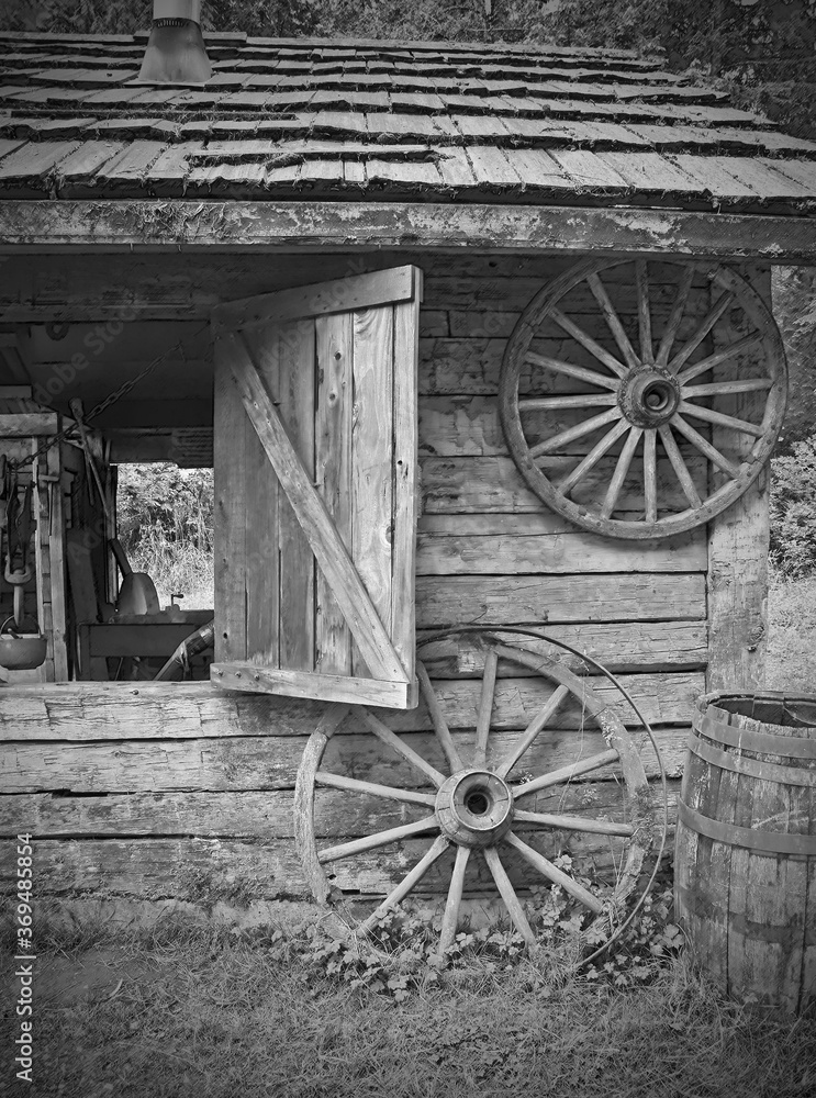 This is an antique scene of old wagon wheels near a log cabin outbuilding with a shutter window open.