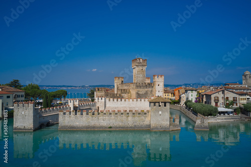 The famous Sirmione Castle. Frontal close aerial view. Reflections of the castle in the water in the background blue sky Sirmione, Lake Garda, Italy.