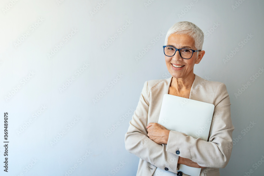 Confident businesswoman with folded arms with laptop standing looking intently at the camera over grey with copy space. Studio portrait of a senior woman posing against a grey background Confident businesswoman with folded arms with laptop standing looking intently at the camera over grey with copy space. Studio portrait of a senior woman posing against a grey background