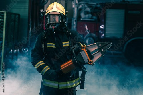 Portrait of a brave firefighter standing confident wearing full protective equipment, turnouts and helmet. Dark background with smoke and blue light. © qunica.com