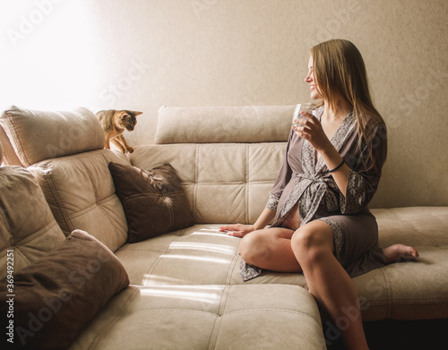 blonde pregnant girl with glass with water in hand is sitting and playing with cat at home on the beige sofa in morning sun lights. lifestyle concept, free space