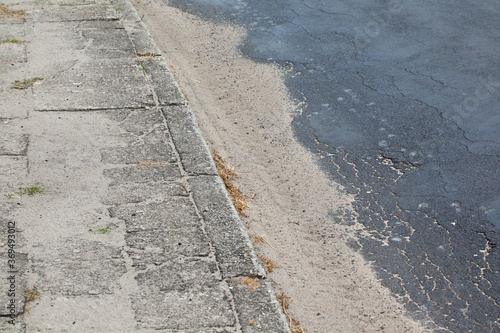 Stary, spękany chodnik z betonowych płyt wraz z krawężnikiem i fragmentem asfaltowej popękanej jezdni.