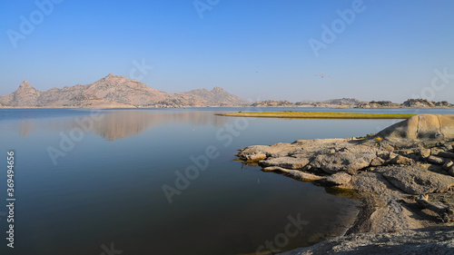 An image of landscape of Jawai dam with clear blue sky and Aravalli mountain ranges with its reflection in water at Jawai in Rajasthan India