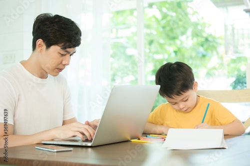 father working with laptop computer while son is drawing, both on the same table.  © Pixpan