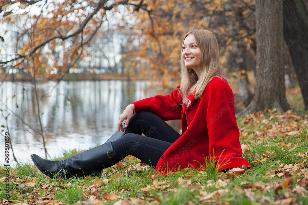 Portrait of a blonde girl in a red coat sitting on the shore of a lake in an autumn park Portrait of a blonde girl in a red coat sitting on the shore of a lake in an autumn park