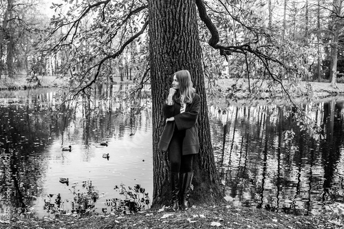 Black and white full-length portrait of a pensive girl in an autumn park next to an old oak tree by the lake
