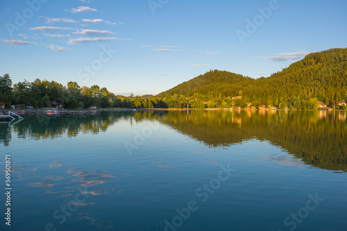 A view of a mountain alpine lake on a evening. Lake with hills, water and blue sky with clouds. Green forest by the lake in reflection in the water beauty in nature