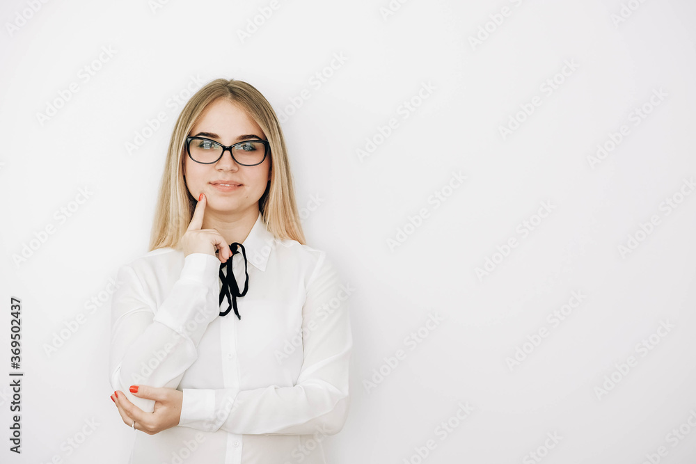 Beautiful blonde girl wears glasses stylish white shirt with a pensive face on a white wall background. Female has clever expression, keeps fore finger near mouth, tries to remember material she Beautiful blonde girl wears glasses stylish white shirt with a pensive face on a white wall background. Female has clever expression, keeps fore finger near mouth, tries to remember material she
