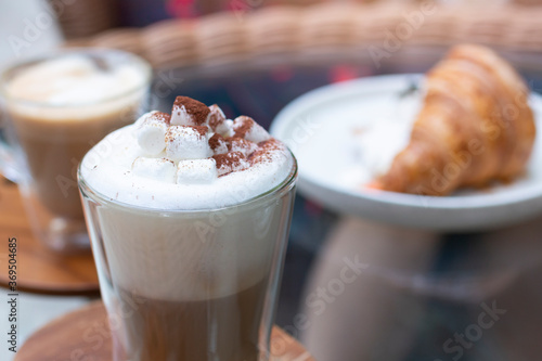 Hot chocolate with marshmallows. Hot Cappuccino in a tall class with Coffee and Croissant on background. Cappuccino or Coffee or Chocolate or Cocoa with marshmallow on top. 