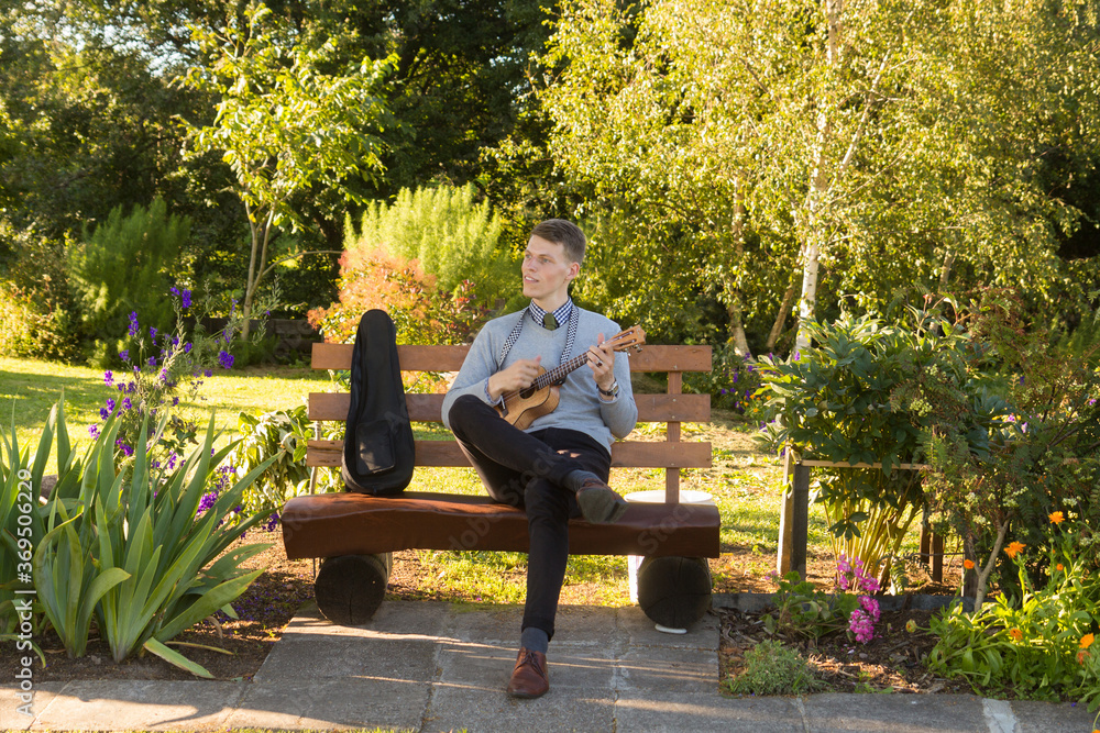 Fototapeta premium Young man sitting on bench in the park and playing ukulele.