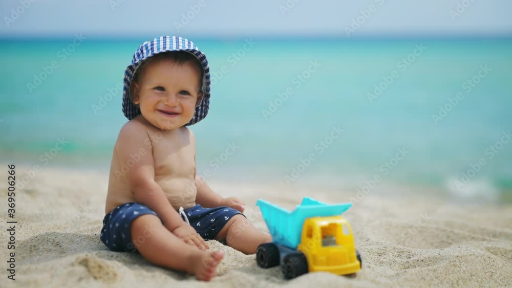 Authentic shot of happy toddler baby boy in blue vichy hat is smiling in camera while playing with a toy truck on a sandy beach during family vacation holiday at sea. Authentic shot of happy toddler baby boy in blue vichy hat is smiling in camera while playing with a toy truck on a sandy beach during family vacation holiday at sea.