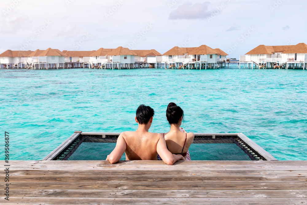 Back of view of romantic couple in love sitting on pier together with beautiful sea. Elegant honeymoon traveler couple hugging on a wooden jetty and enjoy their tropical holiday enjoying ocean view.