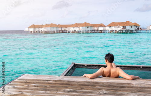 Back view of a young man sitting on a pier with sea and sky in background. Young man on the wooden jetty looking into the ocean. Man on vacation sits on the net by the blue sea looking at the horizon.