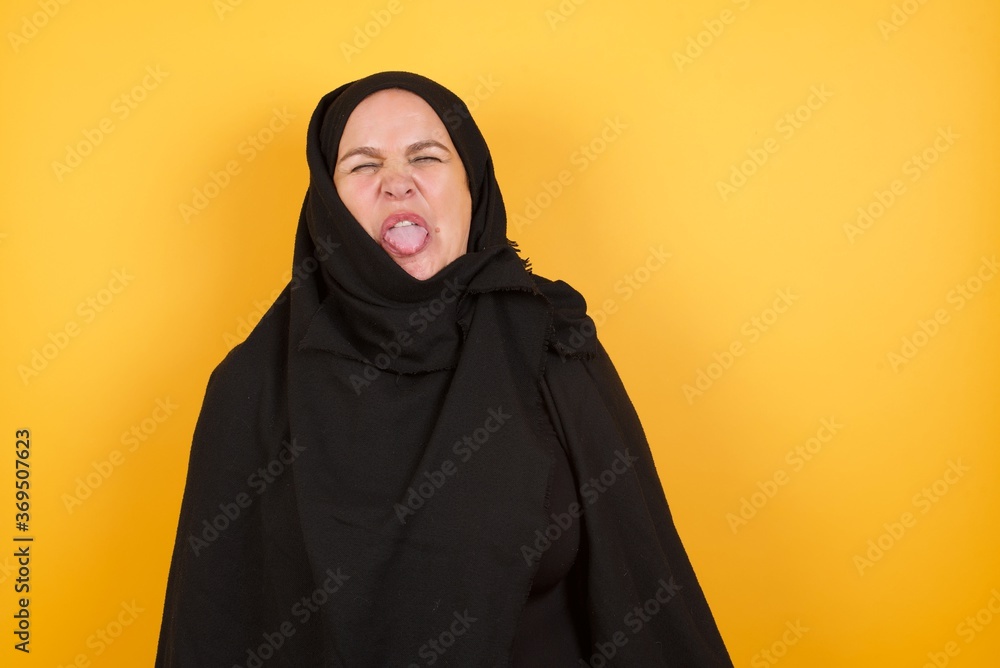Beautiful middle aged muslim woman wearing black hijab over yellow background sticking tongue out happy with funny expression. Emotion concept. Beautiful middle aged muslim woman wearing black hijab over yellow background sticking tongue out happy with funny expression. Emotion concept.