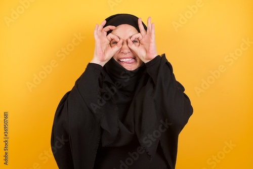 Beautiful middle aged muslim woman wearing black hijab over yellow background doing ok gesture like binoculars sticking tongue out, eyes looking through fingers. Crazy expression. © Roquillo