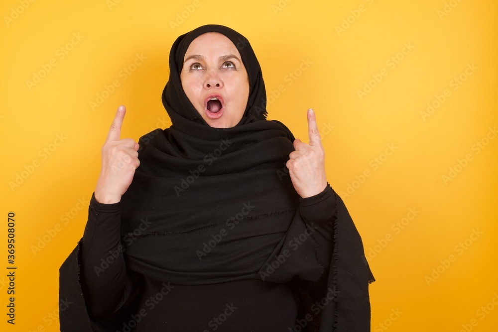 Middle aged muslim woman wearing black hijab over yellow background amazed and surprised looking up and pointing with fingers and raised arms. Middle aged muslim woman wearing black hijab over yellow background amazed and surprised looking up and pointing with fingers and raised arms.