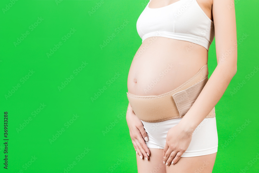 Cropped image of pregnancy belt dressed on pregnant woman in underwear for reducing pain in the back at green background with copy space. Orthopedic abdominal support belt concept Cropped image of pregnancy belt dressed on pregnant woman in underwear for reducing pain in the back at green background with copy space. Orthopedic abdominal support belt concept