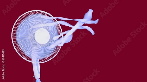 Footage of a white electric fan turning on,blowing cold wind,playing with blue ribbons,floating in the air. Studio shot on red background. Home appliances,modern technology, accommodation concept.