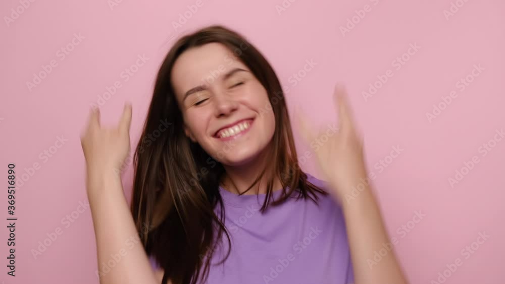 Portrait of cheerful young woman makes rock n roll sign, yells loudly, look at camera, wears purple t-shirt, devil horns hand sign gesture, stands over pink studio background, feels self confident Portrait of cheerful young woman makes rock n roll sign, yells loudly, look at camera, wears purple t-shirt, devil horns hand sign gesture, stands over pink studio background, feels self confident