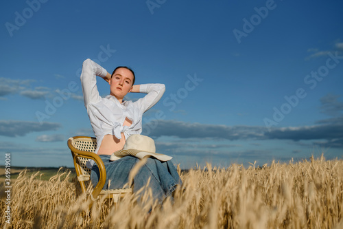 Young woman in casual outfit and a hat is sitting on a vintage chair in the wheat field.