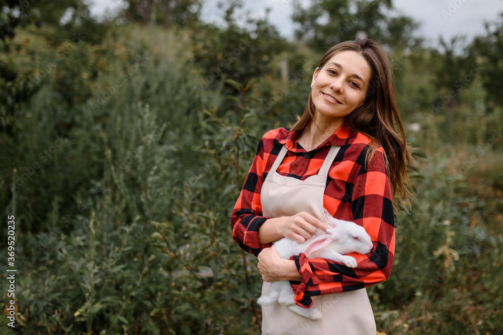 Young happy smiling woman farmer is looking at the camera and holding a white rabbit in her hands outdoors.