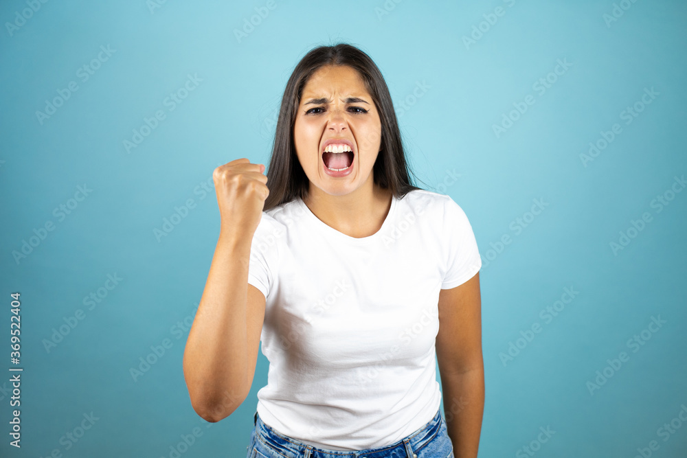 Fototapeta premium Young beautiful woman wearing casual t-shir standing over isolated blue background angry and mad raising fist frustrated and furious while shouting with anger