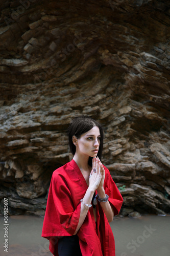Young brunette woman in red kimono keeps her hands folded with a rock in the background