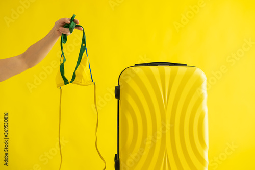 Faceless woman going on a trip holding a bikini next to a suitcase on a yellow background. A female hand is preparing a swimsuit for summer vacation. Copy space.