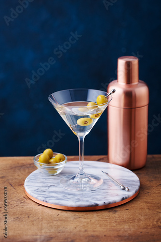 Martini cocktail with green olives, shaker on marble board, wooden table. Copy space.