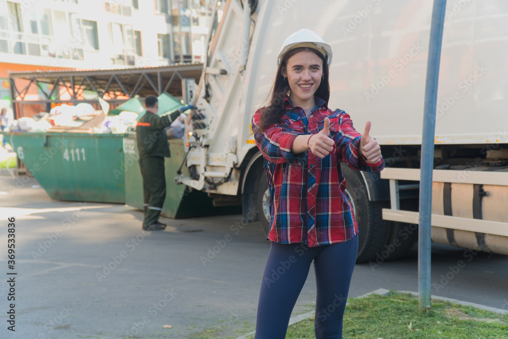 Obraz premium Woman in construction clothes with a white helmet in a plaid shirt on the background of the garbage chute.