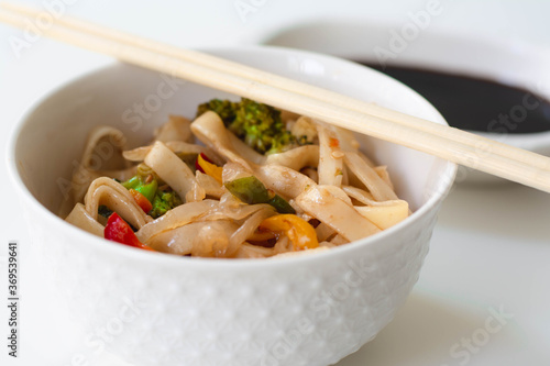 Chinese take out food style egg noodles with broccoli, onions, bell peppers and green beans. White aesthetic, soy sauce and chopsticks in the background.