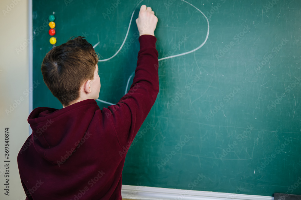 Student boy standing in front of chalkboard in the classroom at school writing. Education concept.