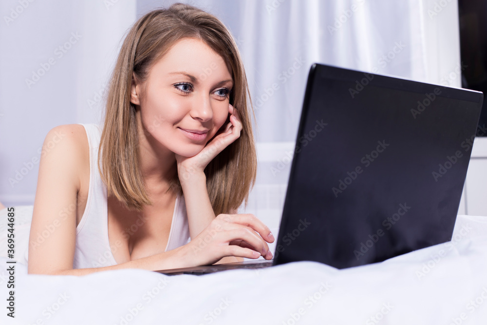 Beautiful blonde woman working on a laptop in her bed in pajamas, smiling Beautiful blonde woman working on a laptop in her bed in pajamas, smiling
