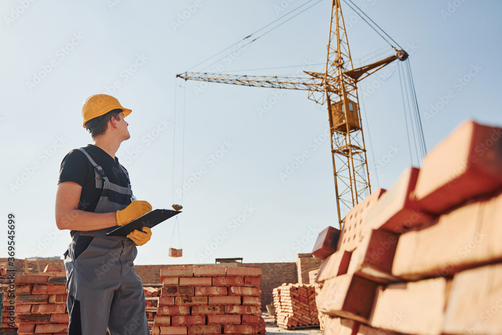 Holds notepad with documents. Construction worker in uniform and safety equipment have job on building
