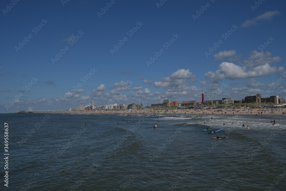 Das Ufer von Den Haag  mit dem roten Leuchtturm, der blaue himmel mit weißen Wolken aus dem Wasser gesehen. Viele Wassersportler schwimmen auf den Wellen des Meeres mit ihren Surfbretter. 
