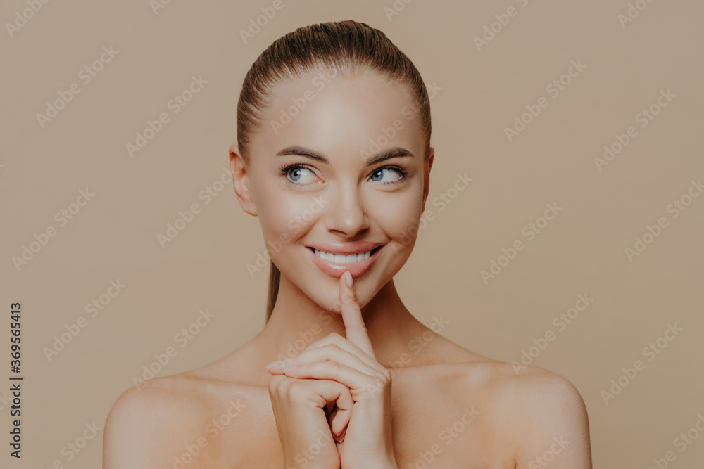 Portrait of good looking woman looks aside mysteriously, keeps finger on chin and smiles broadly, looks away and shares secret of beauty, has perfect glowing skin, naked body, natural makeup Portrait of good looking woman looks aside mysteriously, keeps finger on chin and smiles broadly, looks away and shares secret of beauty, has perfect glowing skin, naked body, natural makeup