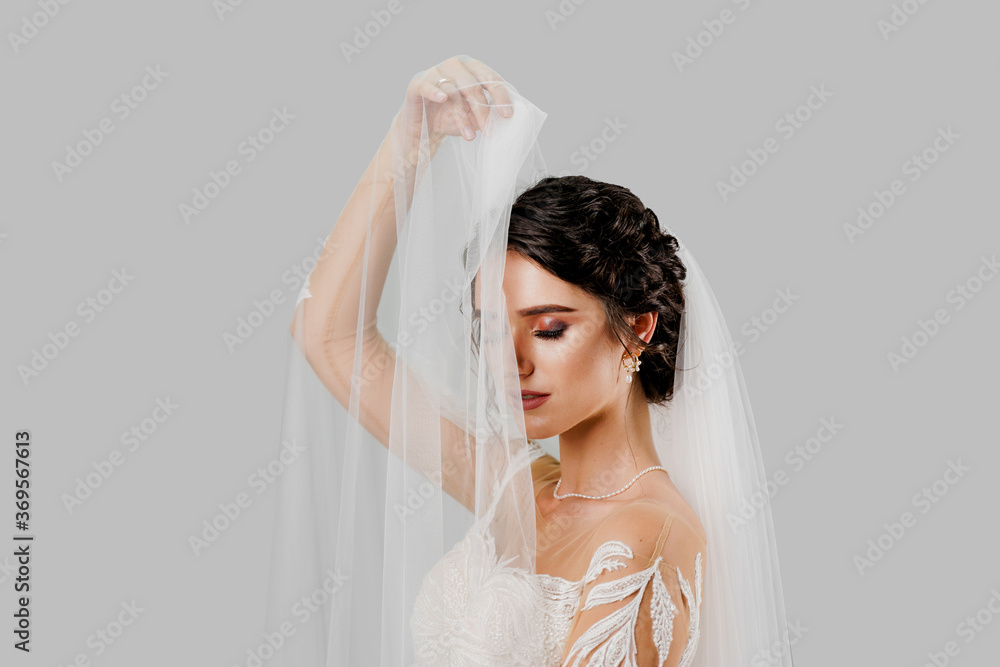 Young attractive bride in wedding dress touches bridal veil on white background in studio. Advert for social networks for wedding agency and bridal salon Young attractive bride in wedding dress touches bridal veil on white background in studio. Advert for social networks for wedding agency and bridal salon