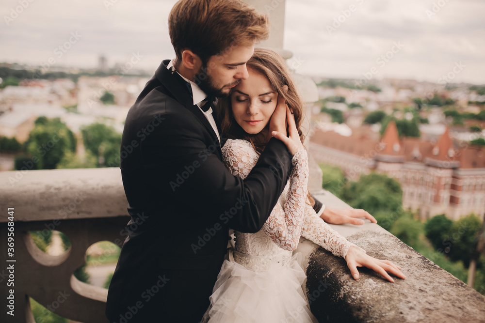 Happy newlywed. beautiful bride and stylish groom are hugging on the balcony of old gothic cathedral with panoramic city views Happy newlywed. beautiful bride and stylish groom are hugging on the balcony of old gothic cathedral with panoramic city views