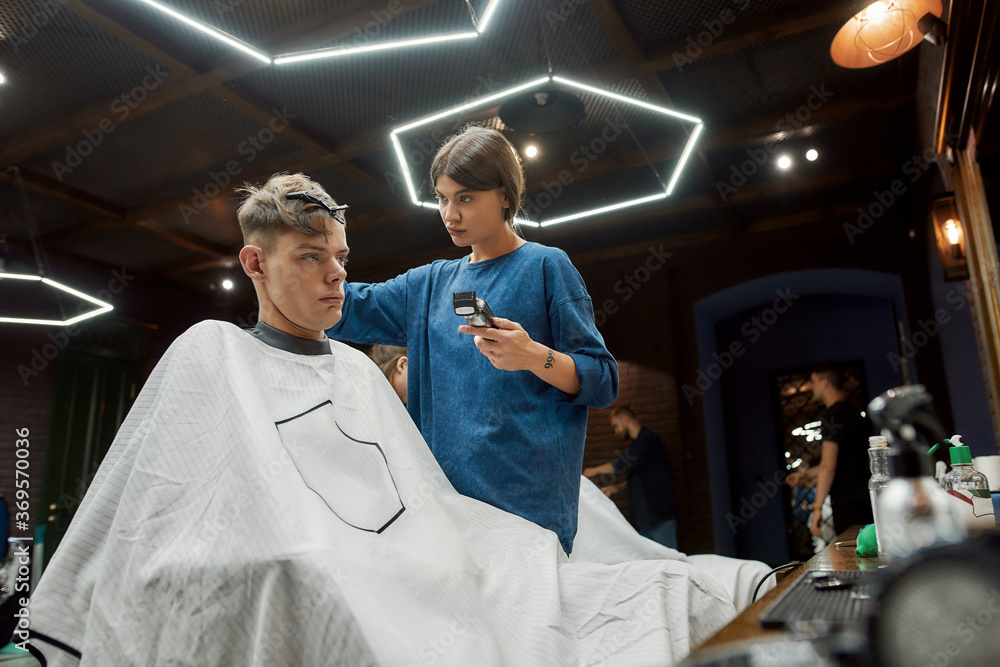 Side view of a young handsome guy sitting in barber shop chair while professional barber girl working with hair clipper, making trendy haircut for him Side view of a young handsome guy sitting in barber shop chair while professional barber girl working with hair clipper, making trendy haircut for him